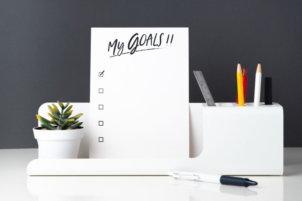 My goals list on notepad at modern office stationery on white table and dark grey wall.pencil, notepad,ruler,pen and pencil box with plant