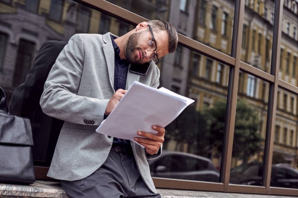 Business communications. Young businessman standing leaning on wall on the city street talking on smartphone with partner reading documents