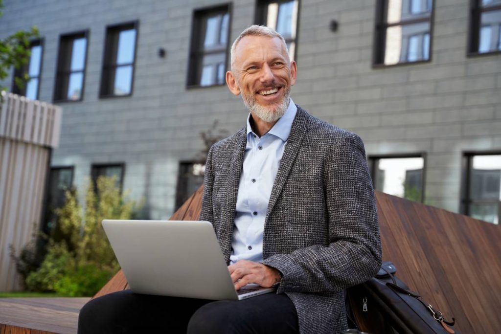 Portrait of cheerful elegant middle aged businessman smiling while working on his laptop, sitting on the bench near modern building outdoors. Business, career concept. Horizontal shot