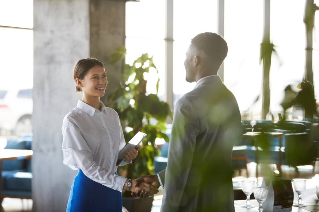 Smiling pretty young business lady with hair bun holding digital table and shaking hand of business partner after signing contract in cafe