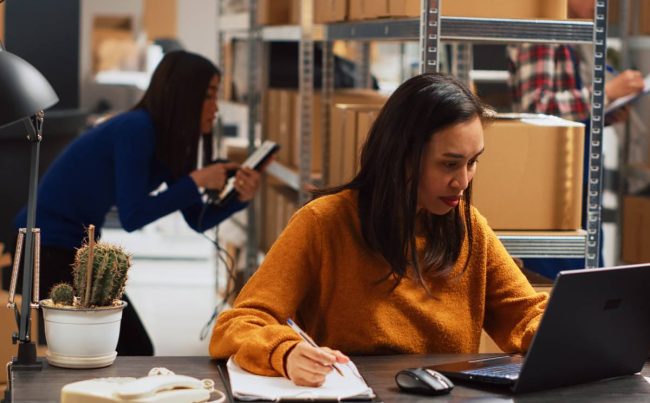 Young woman organizing merchandise in piles, putting retail products in packages for distribution. Female entrepreneur using laptop to check inventory and work on financial planning.