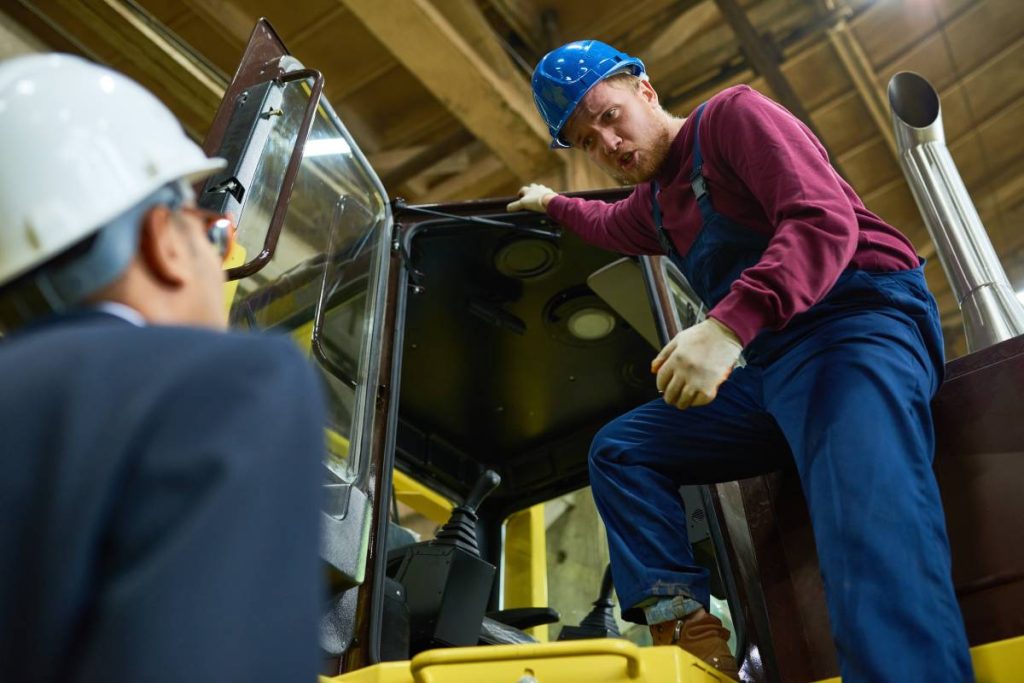 Low angle portrait of worker getting into tractor and looking down at businessman wearing hardhat