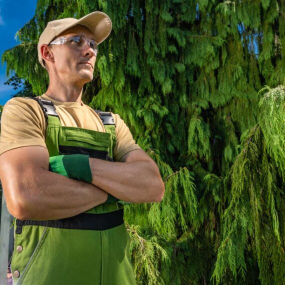 A man in a tan shirt and green overalls stands with his arms crossed. He gazes into the distance while positioned next to a large green tree. The sun is shining, and the setting is a well-kept yard.
